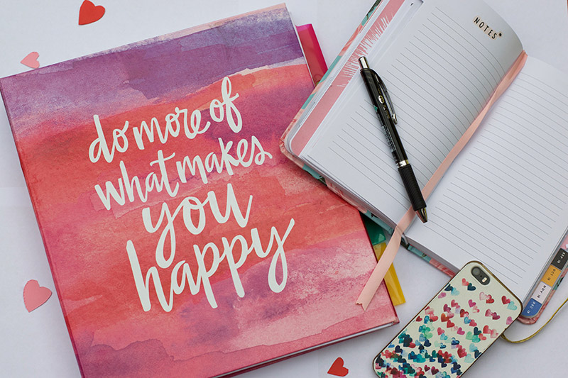 "Do More Of What Makes You Happy" pink folder, with notebook, pen and phone with hearts.